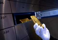 A white-gloved hand holds a shiny gold bar in front of a wide, narrow box that contains several more gold bars.