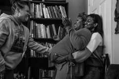 A black-and-white photograph shows three Black women standing in front of a bookshelf. One woman, pregnant, leans her back against another, who reaches around to cradle her baby bump. The third reaches out to touch her belly.