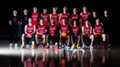 Two rows of young men in basketball uniforms, mostly with red jerseys. The bottom row is seated and reflected in the shiny surface of a black floor.