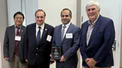 Four men in business suits stand shoulder to shoulder. The third from the left is holding a small plaque.