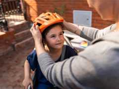 A mother placing an orange bike helmet on her young daughter's head.