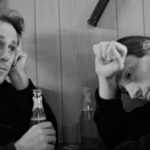 A black-and-white photos of two men looking despondent while holding their head in their respective hand, with the other hand holding a glass bottle of beer.