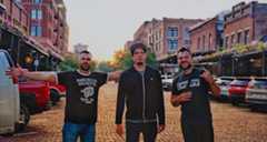 Three men stand in the middle of a cobblestone street with retro small-town strip malls surrounding them and a sun setting behind them.