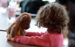 A child with brown curly hair, seen from the back, holds a teddy bear.
