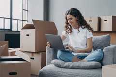 A woman sitting on couch surrounded by moving boxes changing her address online.