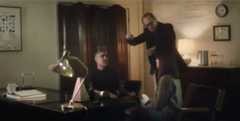 Hayden and Feist sit in an office, while Steve Buscemi stands between them holding a ring on a necklace as if to hypnotize them