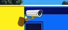 A security camera is mounted on a wall split between yellow and blue, with trees and sky visible above.
