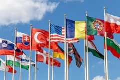 A group of flags from countries from around the world on flagpoles, fluttering in the wind against the backdrop of a blue sky.