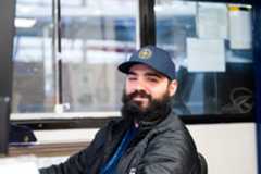 A man with a beard and cap smiles while sitting at a desk in an office, with paperwork and a window in the background.