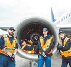 Nolinor-Carriere-Team Four airport workers in safety vests pose near a jet engine, showcasing teamwork and aviation maintenance.