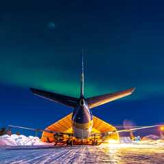 A large airplane is parked in front of a hangar under a night sky illuminated by the northern lights. Snow covers the ground.