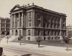 Sepia photo of a large building with columns and stairs. Young trees are seen in the foreground.