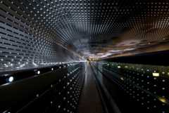 We look through a tunnel lined with dozens of strings of bright white lights nestled in silver-gray slats that zoom away from us in this horizontal photograph. At the far end of the tunnel, at the center of the photograph, the alternating lines of lights and slats almost come together around a narrow, capsule-shaped area of golden yellow light. The flat ceiling curves down to meet the wall to our left. The curve continues to make a C-shape before extending straight down the remaining height of the wall, turning the profile of the open space into a backward-facing P. Some of the lights are bright, some fading, and some are off to create a loose pattern of dark squares floating in a field of light. The lights seem brighter at the far end of the tunnel. The overall impact of the view is a starburst radiating in diagonal lines, coming from a point at the center of the photograph.