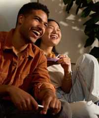 A happy couple laughing while watching TV, sitting on a cozy couch with snacks in hand