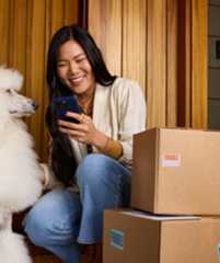 A woman smiling while checking her phone, sitting next to her fluffy white poodle and some fragile-labeled packages