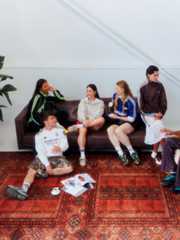 A group of young people, some sitting and some standing, are gathered on a colourful patterned rug in what appears to be a living room or indoor space.
