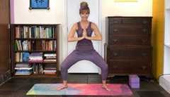 Lorraine Ladish yoga lorraine ladish standing on a yoga mat in her home doing a squatting pose with hands together