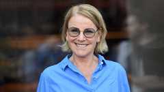 Fotografie zeigt eine lächelnde Frau mit blauer Bluse, blondem Kurzhaarschnitt und Brille.