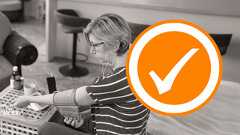 Ein Frau sitzt im Wohnzimmer an einem kleinen Tisch und misst sich ihren Blutdruck mit einem Blutdruckmessgerät.