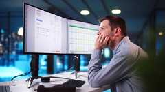 Ein Mann sitzt vor zwei großen Monitoren und betrachtet eine Tabelle in einem Büro bei Abendlicht.