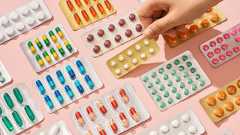 Viele bunte Tabletten‑ und Kapselblister liegen verstreut auf rosa Untergrund, während eine Hand einen goldenen Blister mit weißen Pillen aufhebt.