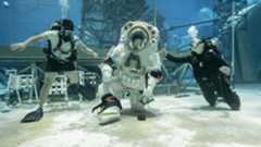 A person wearing a white spacesuit with a clear helmet kneels down to pick up a rock at the bottom of a large sandy pool with two people wearing dark swimsuits on either side of them