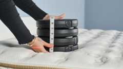 Pressure relief lab test image shows a stack of black weights being placed on the Saatva Classic mattress, in the left hand shoulder area, to measure material indentation and recovery. A read laser line points to the original starting point of the weights so we have a visual record of how much the weight is sinking down, in addition to our two measuring tapes used to record indentation.