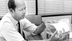 a black and white photo of a man in a white button down shirt and black tie holding a piece of space equipment