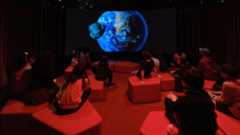 An Earth is projected on a dark screen as children sit on blocks in a dimly red-lit theater