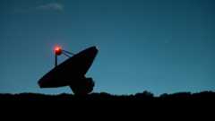 A dark silhouette of a satellite dish tilts to the left next to the dark horizon with a dark blue night sky in the background