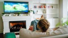 A woman is watching television in a living room, the television is placed above a fire place and she is eating popcorn.
