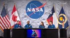 Four people wearing blue jumpsuits sit behind a large table with microphones along with four flags and a circle in the middle of the wall reading "NASA"