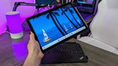 The Lenovo ThinkPad X12 Detachable (Gen 2) held in the hand with the stylus attached, with the detached keyboard lying on a desk in the background.