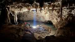 a group of people stand near a shaft of light in a large cave with many stalactites