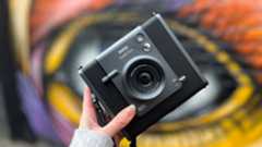 A hand holds up the Instax Wide Evo camera against a colourful graffiti wall
