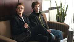A still from the movie The Social Network showing Justin Timberlake and Jesse Eisenberg sitting side-by-side on a brown sofa.