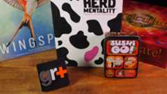 A collection of games (Wingspan, Herd Mentality, Sushi Go, and Articulate) on a wooden table in front of a GR+ logo