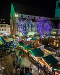 Dieser Alternativtext wurde KI-generiert: Ein nächtliches Bild zeigt einen Weihnachtsmarkt vor einer beleuchteten Kirche. Buden und Bäume sind festlich geschmückt und mit Lichtern versehen.