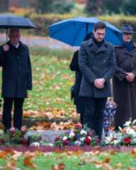Dieser Alternativtext wurde KI-generiert: Mehrere Personen mit Regenschirmen stehen auf einem Rasen vor Blumen und Kränzen. Im Hintergrund sind Bäume und herabgefallenes Laub zu sehen.