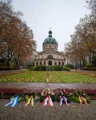 Dieser Alternativtext wurde KI-generiert: Ein Gebäude mit einer Kuppel steht inmitten von Bäumen mit herbstlichem Laub. Im Vordergrund befinden sich Blumen- und Kranzarrangements.
