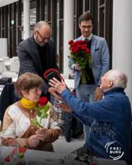 Dieser Alternativtext wurde KI-generiert: Drei Personen stehen in einem hellen Innenraum. Eine Person übergibt eine rote Rose an eine andere, während eine dritte einen Blumenstrauß hält.