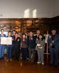 Dieser Alternativtext wurde KI-generiert: Eine Gruppe von Menschen steht in einem Raum mit Holztäfelung zusammen und hält Dokumente und Zeichen in der Hand. Das Bild ist farbig und zeigt ein formelles Arrangement.