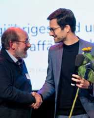 Dieser Alternativtext wurde KI-generiert: Zwei Personen stehen sich gegenüber und geben sich die Hand. Im Hintergrund ist eine verschwommene Wand mit Text und eine einzelne Sonnenblume zu sehen.