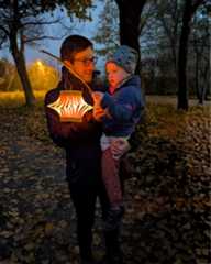 Dieser Alternativtext wurde KI-generiert: Eine Person hält ein Kind auf dem Arm und präsentiert eine Laterne. Im Hintergrund sind Bäume und eine beleuchtete Straße zu sehen.