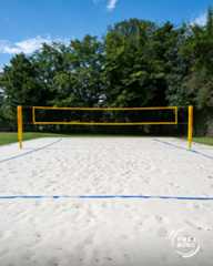 Dieser Alternativtext wurde KI-generiert: Ein Volleyballfeld mit gelben Pfosten und Netz befindet sich auf einer Sandfläche. Im Hintergrund sind grüne Bäume und ein blauer Himmel zu sehen.