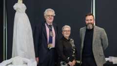 A white embroidered dress is on a mannequin in the background, to the left, and there people stand side by side, smartly dressed