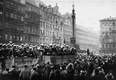 Truckloads of standing uniformed troops drive through a large crowd gathered in the street. The crowd is a combination of civilians and soldiers.