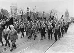 Hundreds of uniformed individuals parade down a street with Adolf Hitler and other Nazi officials leading the group. A few individuals carry flags with swastikas.