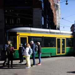 A green-and-yellow tram moves on tracks alongside a group of pedestrians in a city setting under a bright sky.