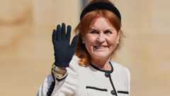 Sarah Ferguson waves her black-gloved hand while wearing a black headband, a cream jacket with a black trim, and large pearl earrings.